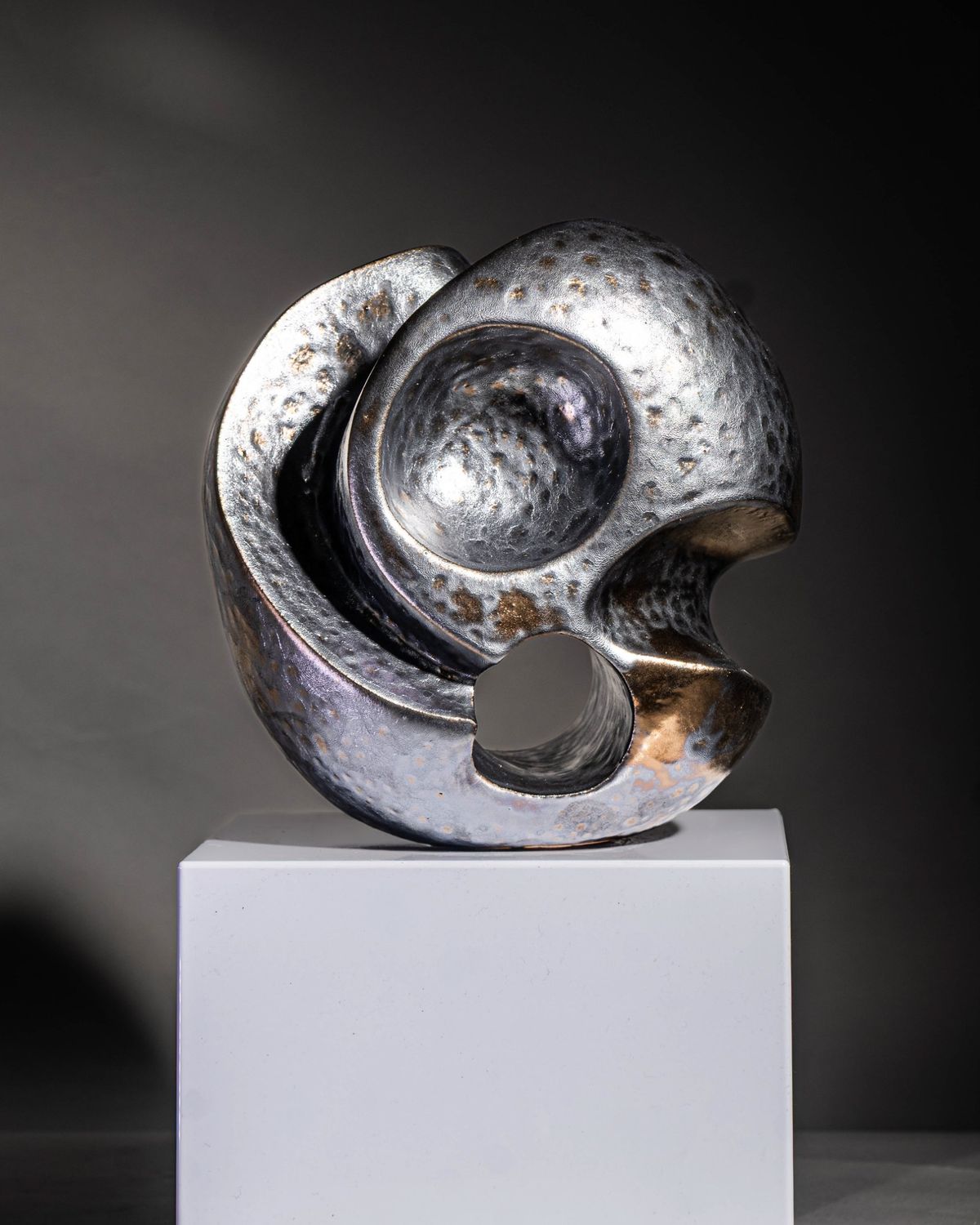 Silver and bronze glazed sculptural form with an open void, on a white plinth