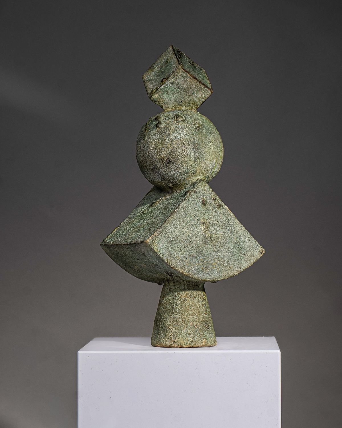Pale green textured sculpture stacking a cube, sphere and wedge on a tapered base